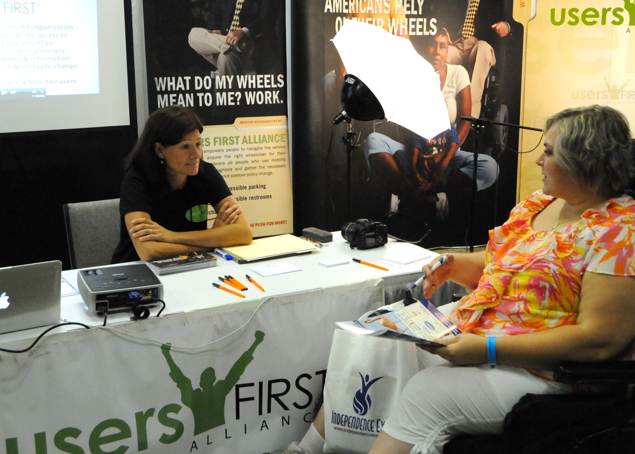 A wheelchair user speaks with a UsersFirst representative at a conference information booth.