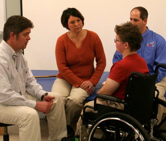 A wheelchair team comprised of a doctor, clinician, and supplier, all sit together speaking with a client.