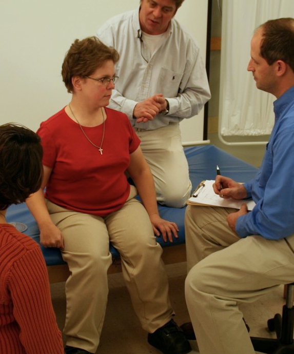 A woman sits on a mat at her Wheelchair Evaluation and Assessment surrounded by her wheelchair team and speaking with her medical equipment supplier who is taking notes on their conversation.