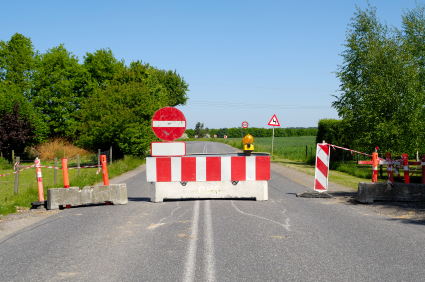 A warning sign and cement barrier block traffic from traveling down a road.