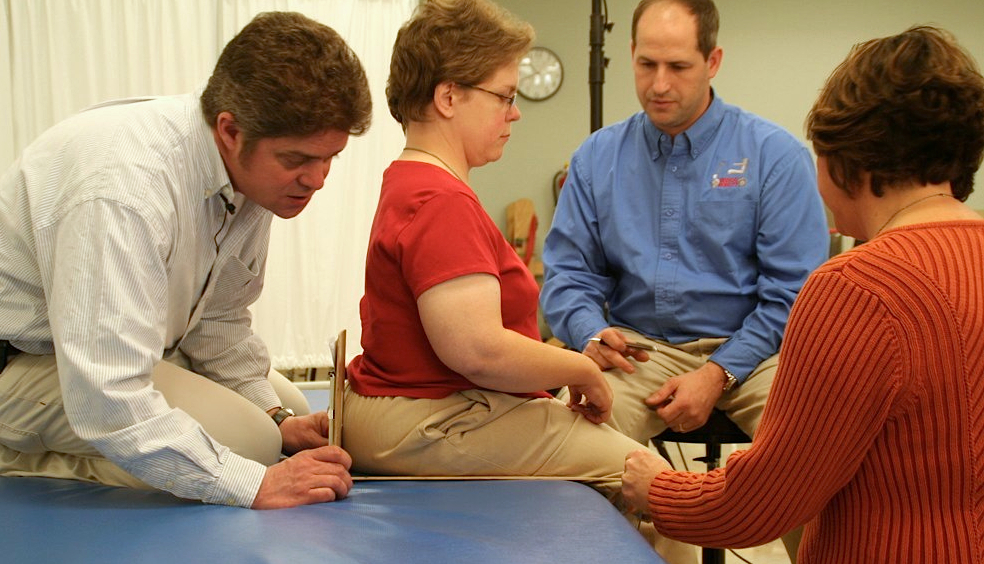 A wheelchair user seated on a cushioned table is measured by two clinicians while a medical equipment supplier takes notes.