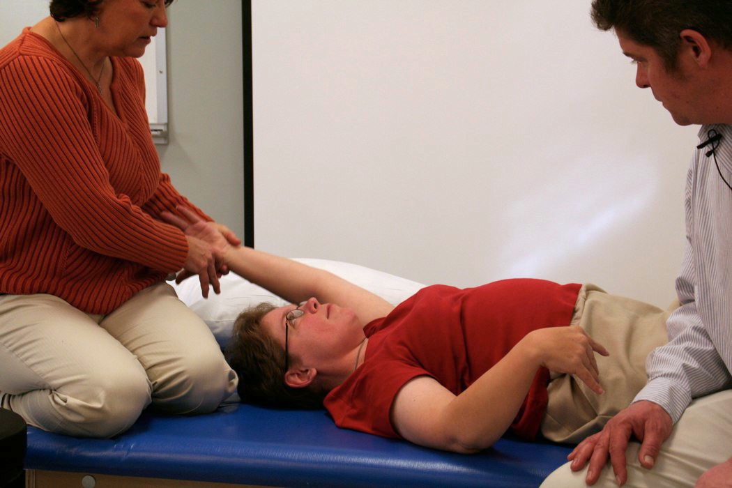A woman lays on a soft mat while a clinician guides her arm up and over her head as a part of her wheelchair evaluation and assessment. 