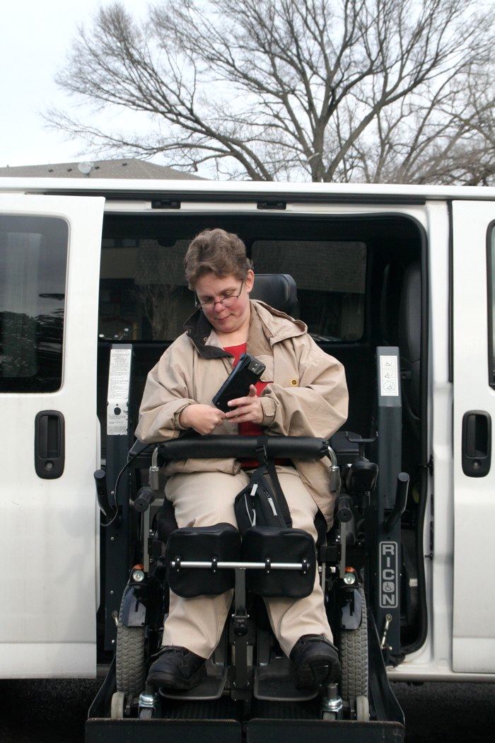 A woman exits her van by lowering herself on her electronic wheelchair lift.